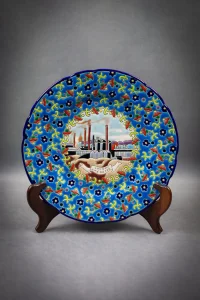 Assiette en émaux de Longwy polychromes à décor de fleurs entourant un médaillon central représentant la cité du fer. Assiette sur un présentoir, de face, format portrait.