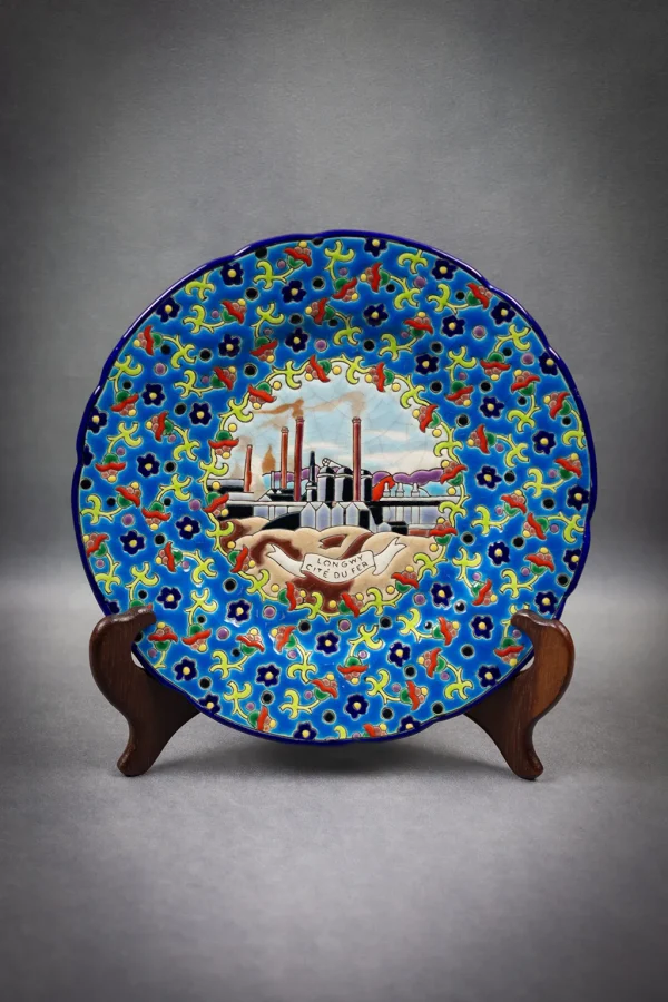 Assiette en émaux de Longwy polychromes à décor de fleurs entourant un médaillon central représentant la cité du fer. Assiette sur un présentoir, de face, format portrait.